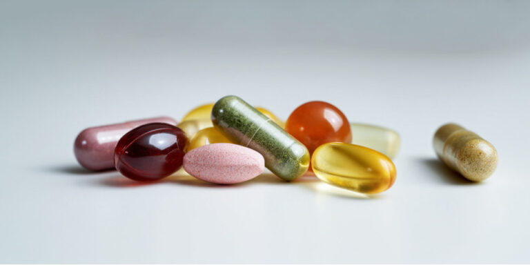 A variety of colourful pills and capsules, including oval, round, and oblong shapes, are scattered on a plain white surface. UK