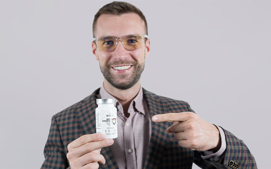 A smiling man wearing glasses and a checked blazer holds up and points to a white bottle of supplements or medication against a plain light grey background. UK