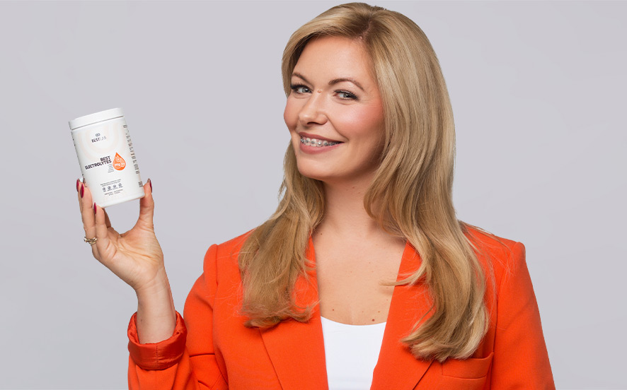 A woman with long blonde hair, wearing an orange blazer and white top, smiles whilst holding up a white container with a label and barcode against a plain light grey background. UK
