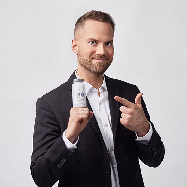 A man in a black suit and white shirt smiles whilst holding a white bottle and pointing at it with his other hand, standing against a plain light background. UK