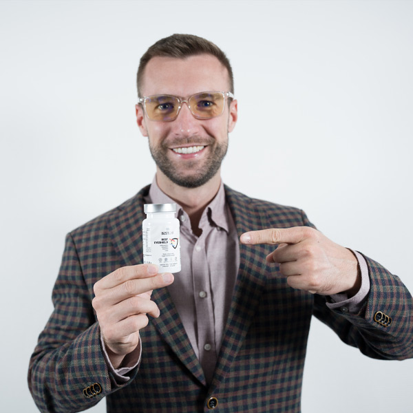 A smiling man in glasses and a checked blazer holds and points to a white pill bottle, standing against a plain white background. UK