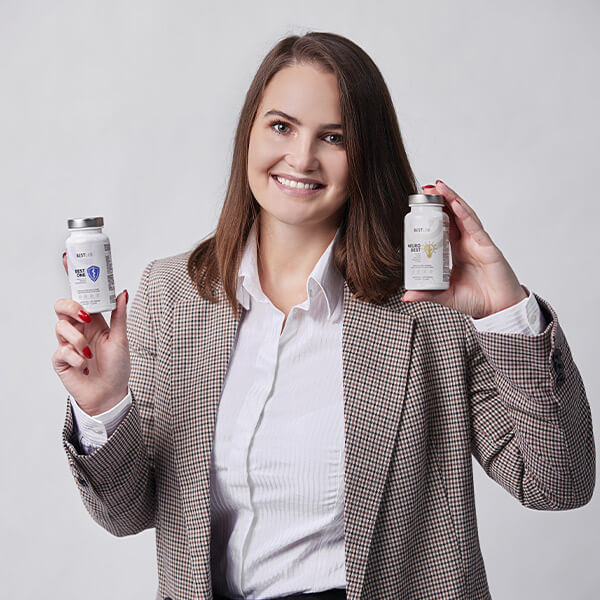 A smiling woman in a checked blazer and white shirt holds up two supplement bottles, one in each hand, against a plain light background. UK