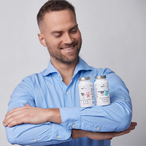 A smiling man in a light blue shirt balances two supplement bottles on his folded arm against a plain light background. UK