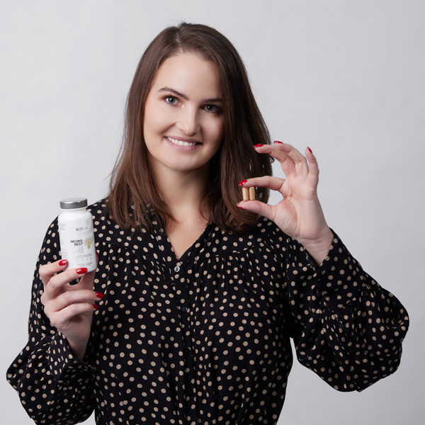 A smiling woman with brown hair in a black polka dot blouse holds a white supplement bottle in one hand and shows a pill with the other hand against a plain white background. UK