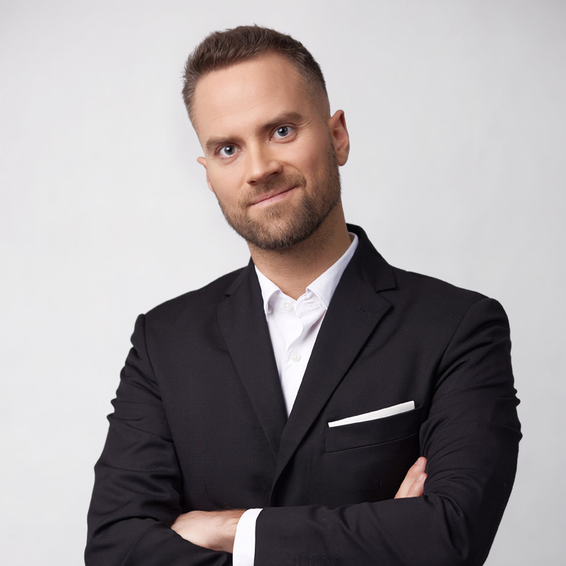 A man with short hair and a trimmed beard, wearing a black suit and white shirt, stands with arms crossed and smiles slightly against a plain light background. UK
