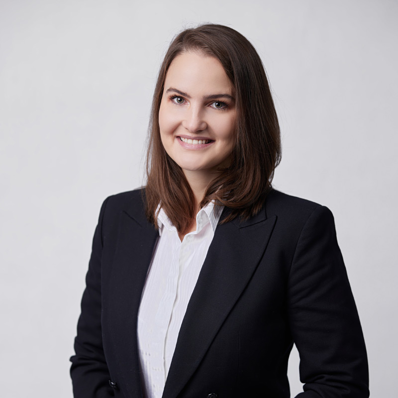 A woman with straight brown hair, wearing a black blazer and white shirt, smiles at the camera against a plain light background. UK