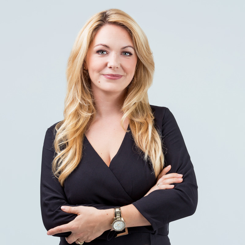 A confident woman with long blonde hair wearing a black dress and a wristwatch stands with her arms crossed, smiling slightly against a light grey background. UK