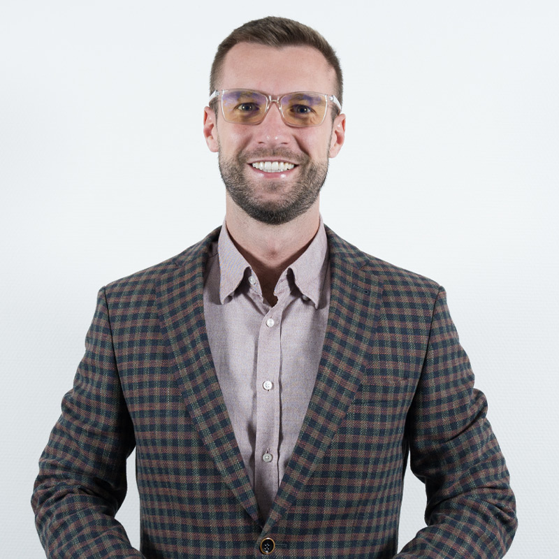 A man wearing yellow-tinted glasses, a checked blazer, and a button-up shirt stands against a plain white background, smiling at the camera. UK