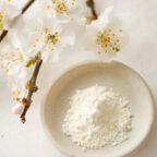 A small white plate holding a mound of fine white powder sits beside delicate white blossoming branches on a light surface. UK