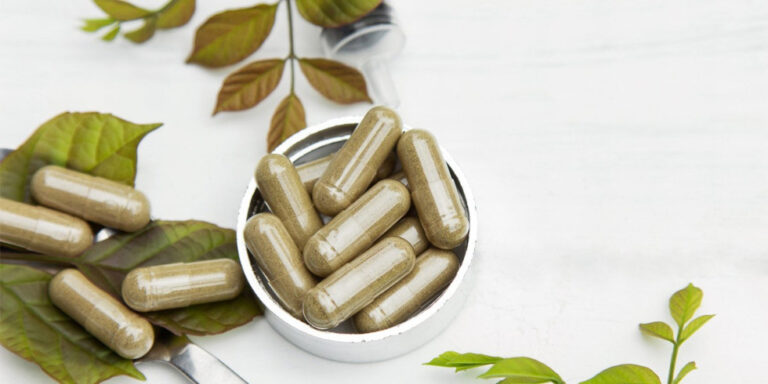 a close up of several brown herbal capsules in a small round container, surrounded by fresh green leaves on a white surface. uk