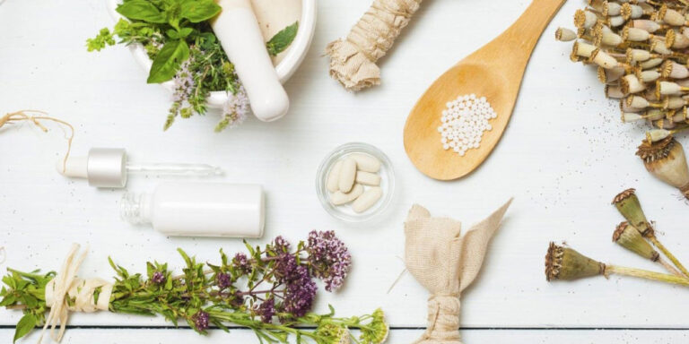 a flat lay of herbal remedies: fresh and dried herbs, a mortar and pestle, a wooden spoon with small white pills, a dropper bottle, a bowl with tablets, and bundles of dried plants on a white surface. uk