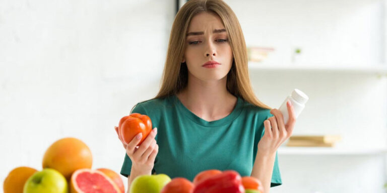 a young woman in a green shirt looks uncertain while holding a tomato in one hand and a bottle of tablets in the other, with a variety of fruit and vegetables on the table in front of her. uk