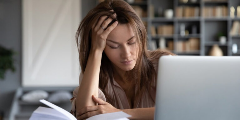 a young woman sits at a desk with her head resting on her hand, looking tired and stressed. she is surrounded by an open book and a laptop, with shelves of books in the background. uk
