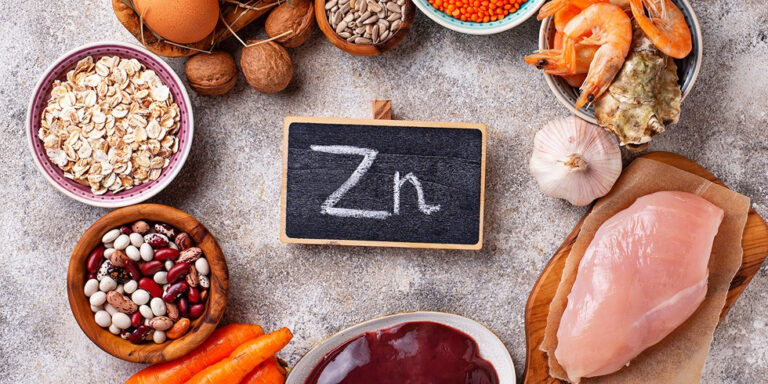 a variety of zinc rich foods, including oysters, prawns, chicken breast, beans, nuts, seeds, eggs, carrots, oats, and garlic, are arranged around a small blackboard with zn written on it. uk