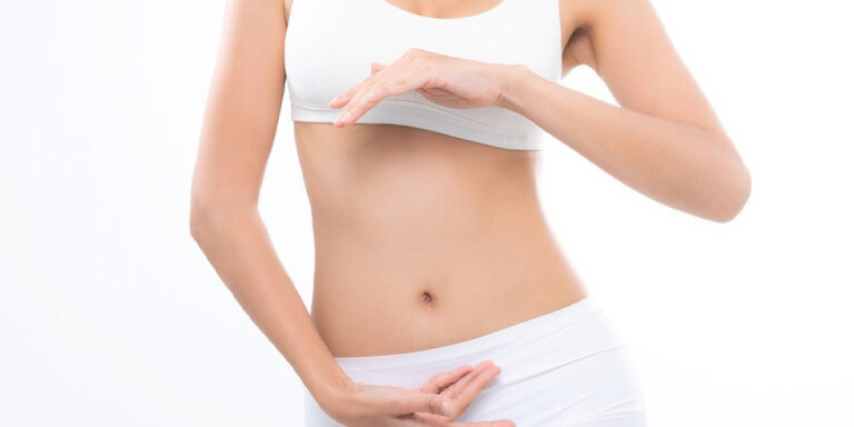 a woman in white underwear and a white sports bra stands against a white background, showing her midriff. she forms a circular shape with her hands near her abdomen, drawing attention to her stomach area. uk