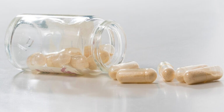 a glass jar tipped on its side spills several beige capsules onto a white surface. some capsules remain inside the jar, whilst others are scattered nearby. uk