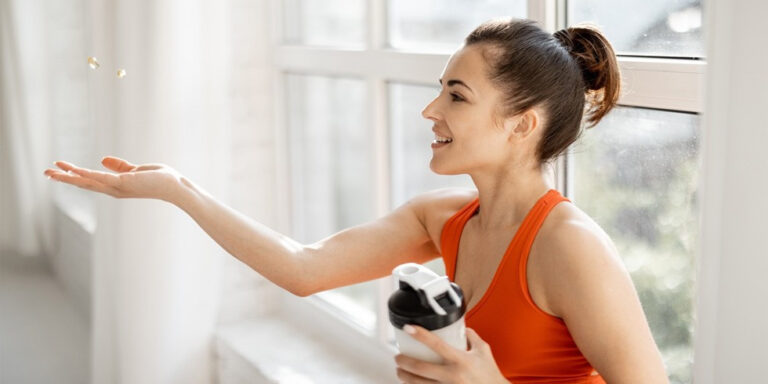 a woman in an orange vest sits by a large window, smiling as she tosses vitamins into the air with one hand while holding a shaker bottle in the other. sunlight streams through the window. uk
