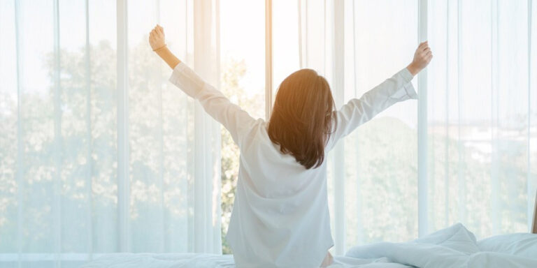 a person with shoulder length hair sits on a bed facing large windows, stretching their arms upwards. soft sunlight filters through sheer curtains, creating a calm and peaceful morning atmosphere. uk
