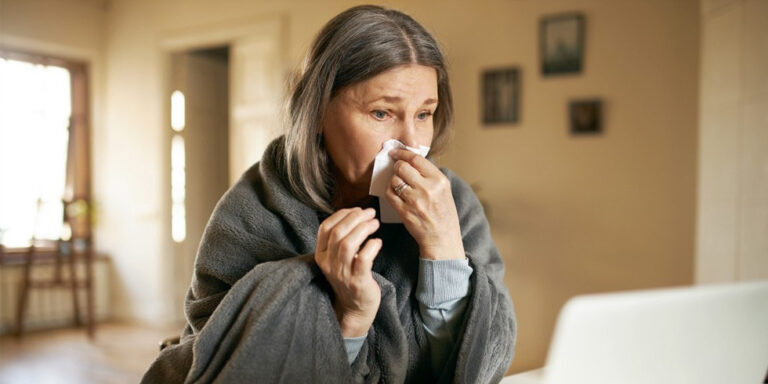 a woman wrapped in a grey blanket sits indoors, holding a tissue to her nose as if she is sneezing or has a cold. she appears unwell and is looking at a laptop in front of her. uk
