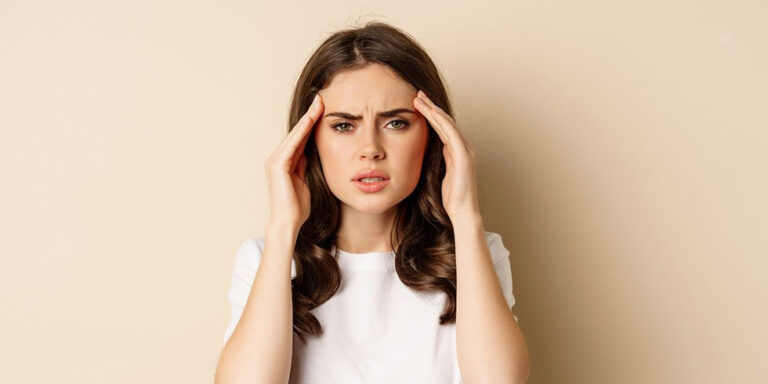 a young woman with long brown hair in a white shirt stands against a beige background, holding her hands to her temples and looking distressed or stressed. uk