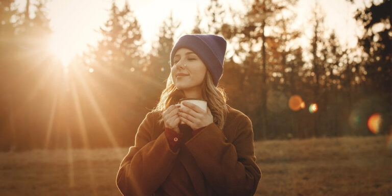 a woman in a brown coat and blue beanie stands outdoors, holding a cup and smiling with her eyes closed, enjoying the sunlight. pine trees and the sun are visible in the background. uk