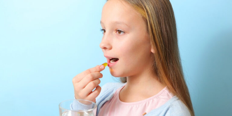 a young girl with long light brown hair holds a glass of water in one hand and is about to take a yellow capsule with the other hand, standing against a light blue background. uk