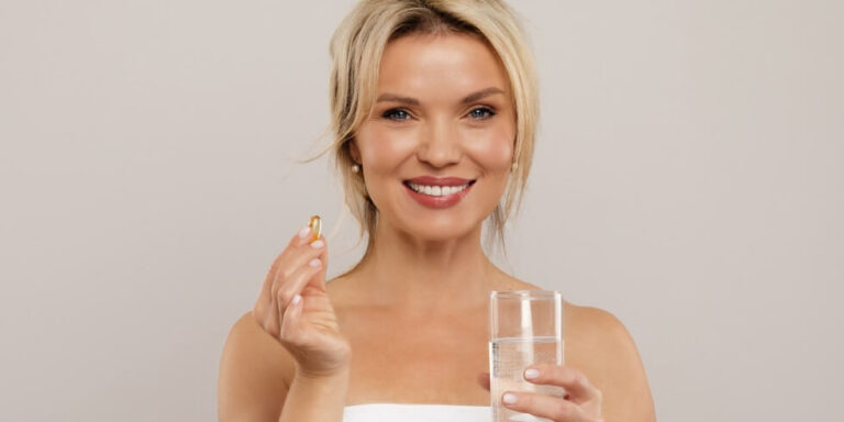 smiling woman with blonde hair holding a yellow capsule in one hand and a glass of water in the other, standing against a plain light background. uk