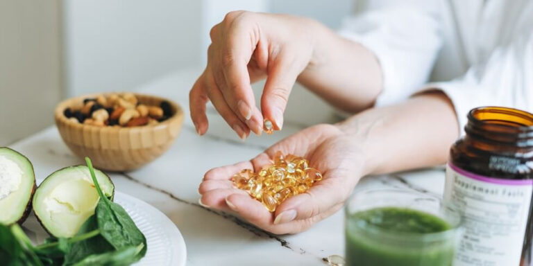 a person holds several yellow supplement capsules in one hand and picks one up with the other. nearby are a bowl of nuts, sliced avocado, leafy greens, and an open supplement bottle on a white table. uk