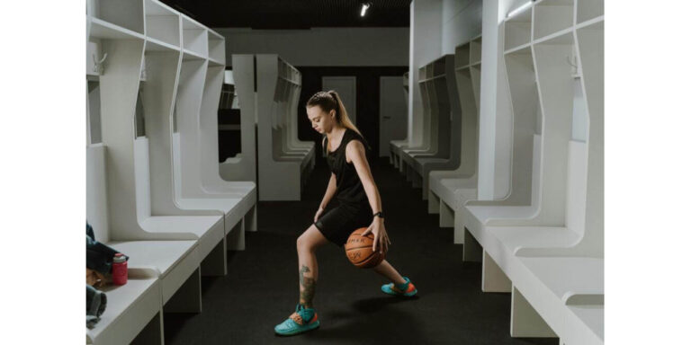 a woman in sportswear dribbles a basketball while practising in an empty changing room with white benches and open lockers on both sides. uk