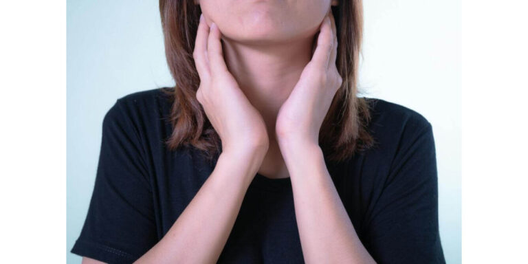a person wearing a black shirt touches both sides of their neck with their hands, possibly indicating discomfort or pain in the throat or neck area. uk