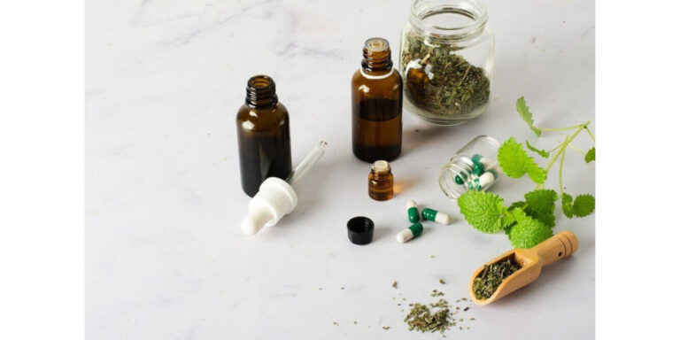 brown glass dropper bottles, capsules, and jars filled with dried herbs are arranged on a white surface, alongside fresh green leaves and a wooden scoop containing dried herbs. uk