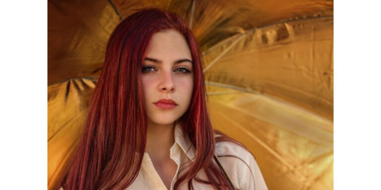 a young woman with long red hair and a neutral expression stands in front of a bright yellow or gold umbrella, wearing a light coloured shirt. the umbrella creates a warm, glowing background. uk