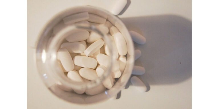 a top down view of a clear bottle filled with white oval tablets, some of which are spilling out onto a white surface. uk