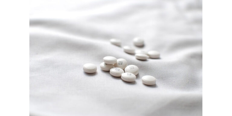 a small pile of white round tablets scattered on a soft, white fabric surface with gentle folds. the tablets have faint numbers and letters imprinted on them. uk