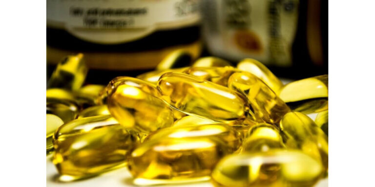 close up of several yellow, translucent gel capsules, likely fish oil or vitamin supplements, with blurred bottles in the background. uk