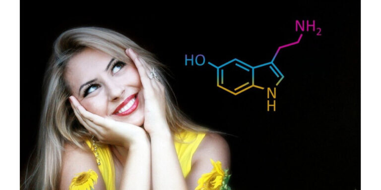 a smiling woman in a yellow top rests her face in her hands and looks upwards. next to her is a colourful chemical structure diagram on a black background. uk