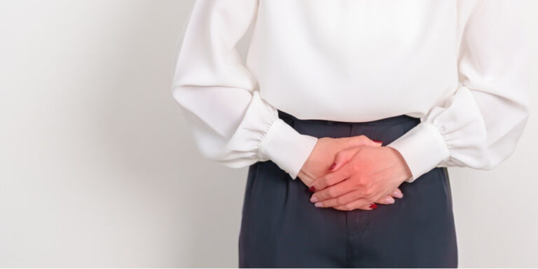 a person in a white blouse and dark trousers is standing with hands clasped over their lower abdomen, suggesting discomfort or pain in that area. uk