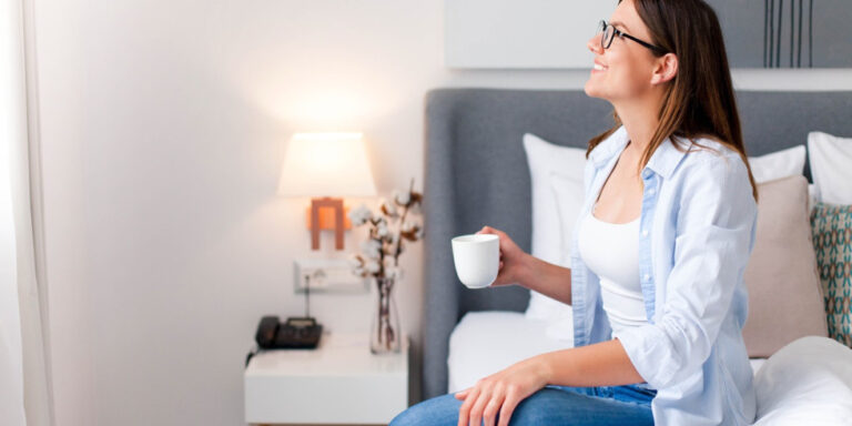 a woman with glasses sits on a bed, smiling and holding a white mug. she wears a light blue shirt and jeans. a bedside table with a lamp, a phone, and decorative items is in the background. uk
