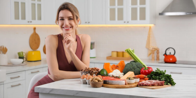 a woman smiles in a modern kitchen, sitting at a worktop with a variety of fresh foods, including vegetables, salmon, cheese, nuts, and meat, arranged in front of her. uk