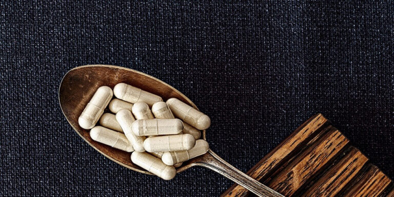 a metal spoon filled with white capsules rests on a dark textured surface next to a piece of wood. uk