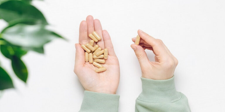a person wearing a light green jumper holds several beige capsules in one hand and picks up one capsule with the other, with a leafy green plant partially visible on the left. uk