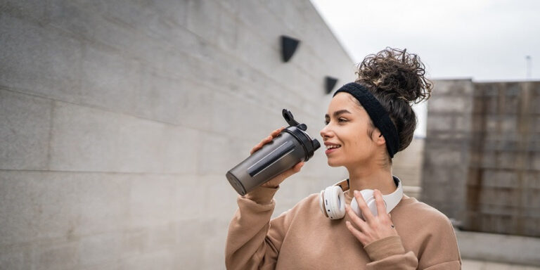 a young woman wearing a headband and headphones around her neck drinks from a sports bottle outdoors. she is dressed in a beige sweatshirt and stands against a grey concrete wall. uk
