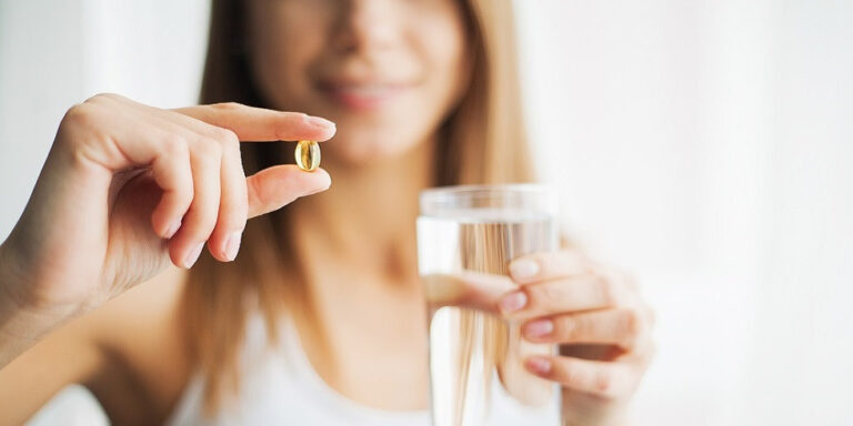 a woman holds a glass of water in one hand and a yellow capsule pill between her fingers in the other, with a blurred background. uk