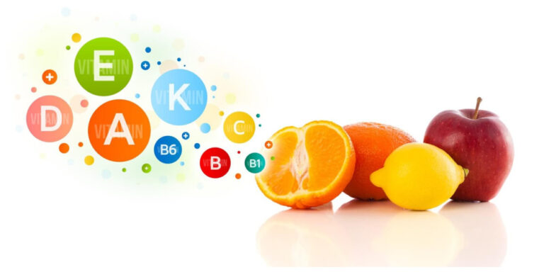 colourful vitamin icons (a, b6, c, d, e, k) float beside fresh fruit: a halved orange, a whole orange, a lemon, and a red apple, suggesting the fruit are sources of various vitamins. uk