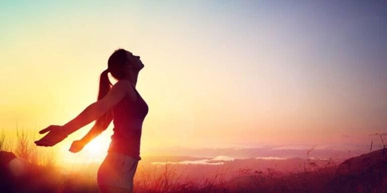 a woman stands outdoors at sunrise or sunset with arms outstretched and head tilted back, silhouetted against a colourful sky, expressing freedom and joy in nature. uk