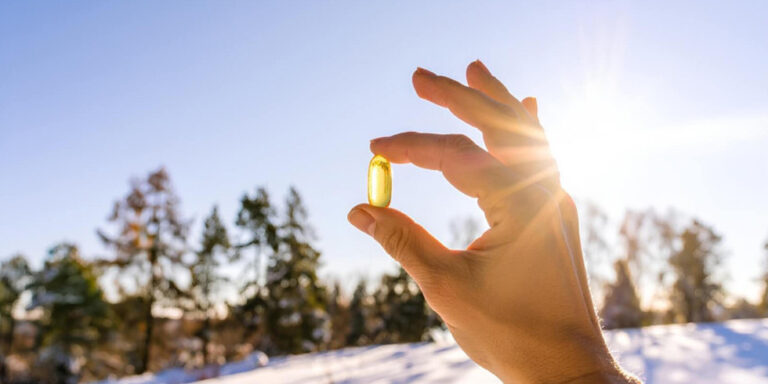 a hand holds a yellow capsule up to the sunlight outdoors, with a snowy landscape and trees in the background under a clear blue sky. uk