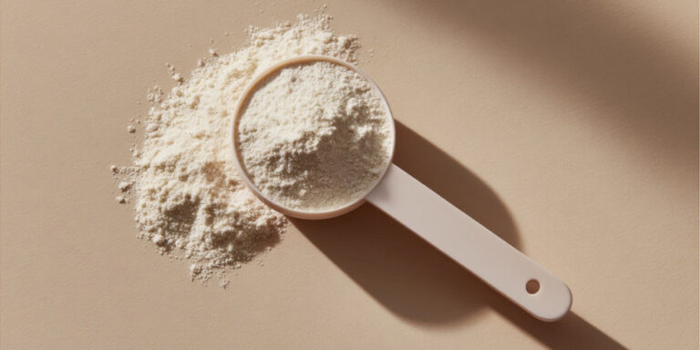 a white plastic measuring scoop filled with a fine white powder, with additional powder spilt onto a smooth beige surface, photographed in bright, natural light. uk