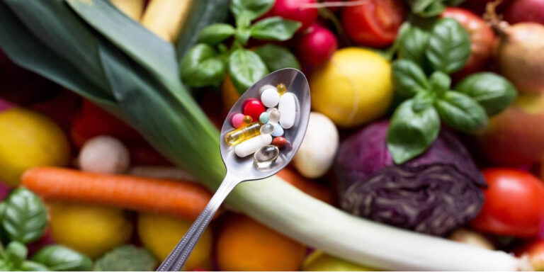 a spoon holding various pills and capsules is held above a colourful assortment of fresh vegetables and herbs, including carrots, cabbage, basil, and radishes. uk