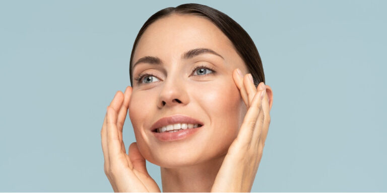 a woman with clear skin and dark hair pulled back gently touches her face with both hands, smiling and looking upwards against a light blue background. uk