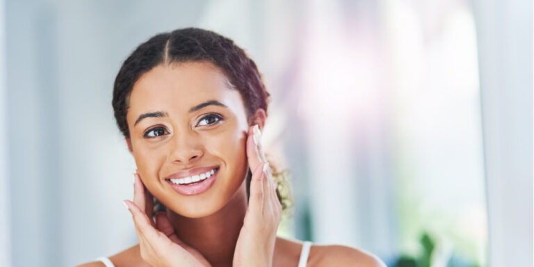 a young woman with curly hair smiles whilst gently touching her face with both hands. she appears happy and confident, standing in a brightly lit room with a soft focus background. uk